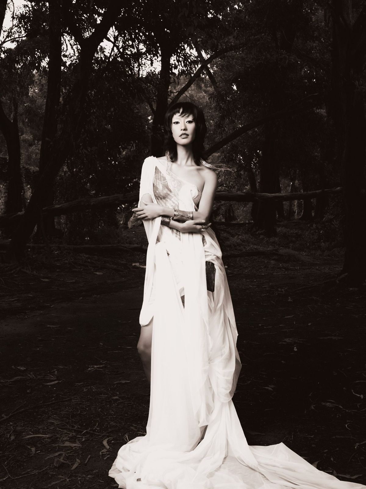 Model in a white draped gown standing in a dark forest